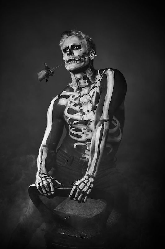 A man with a skeleton painted on his body is holding a rose in his mouth.