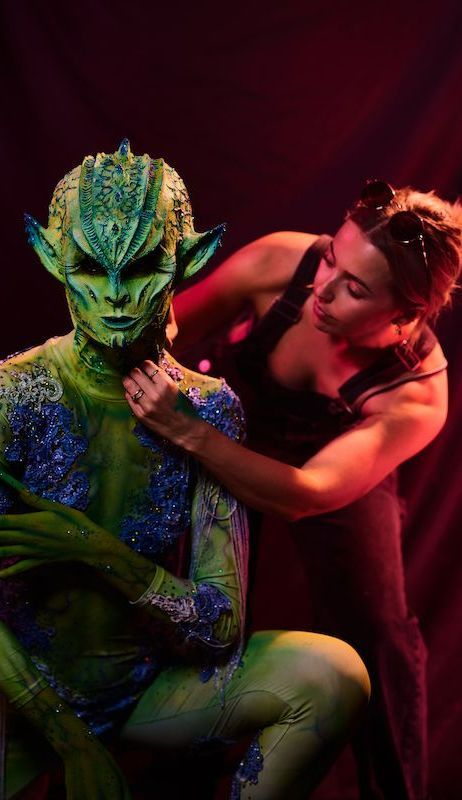 A woman is painting a person 's body with green and blue paint.