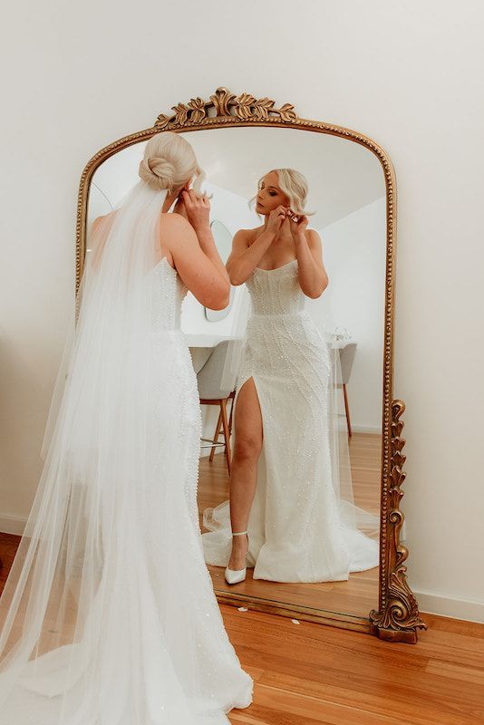 A bride is putting on her earrings in front of a large mirror.