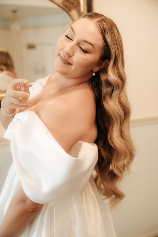 A woman in a wedding dress is applying perfume to her neck.