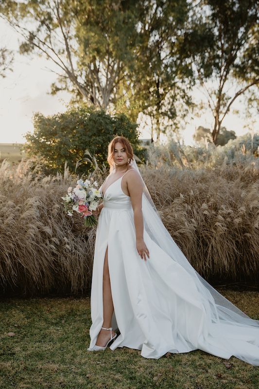 A woman in a white wedding dress is standing in the grass holding a bouquet of flowers.