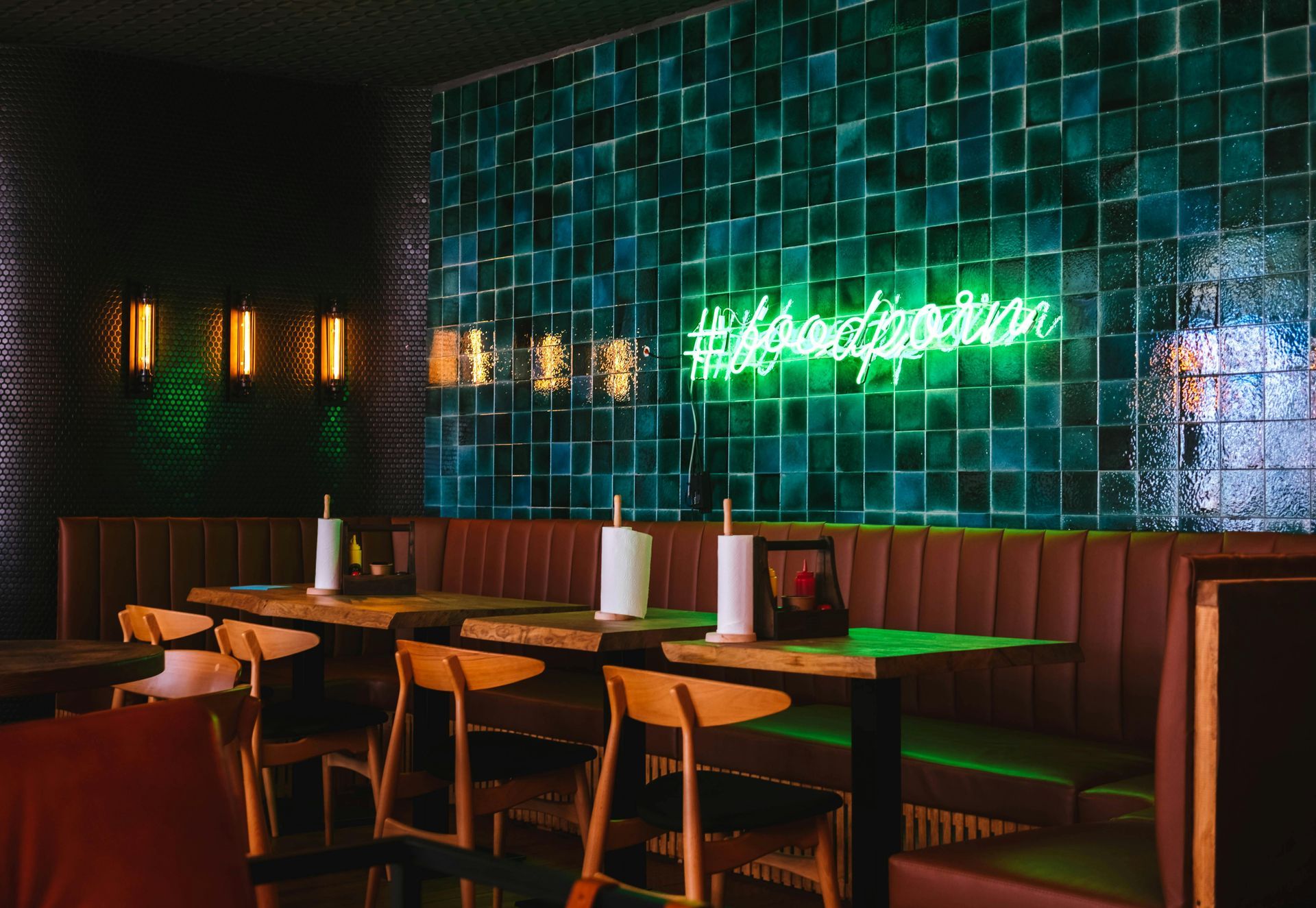 Restaurant interior: booths, tables with paper towels, wooden chairs, teal tile wall, neon sign, warm lighting.