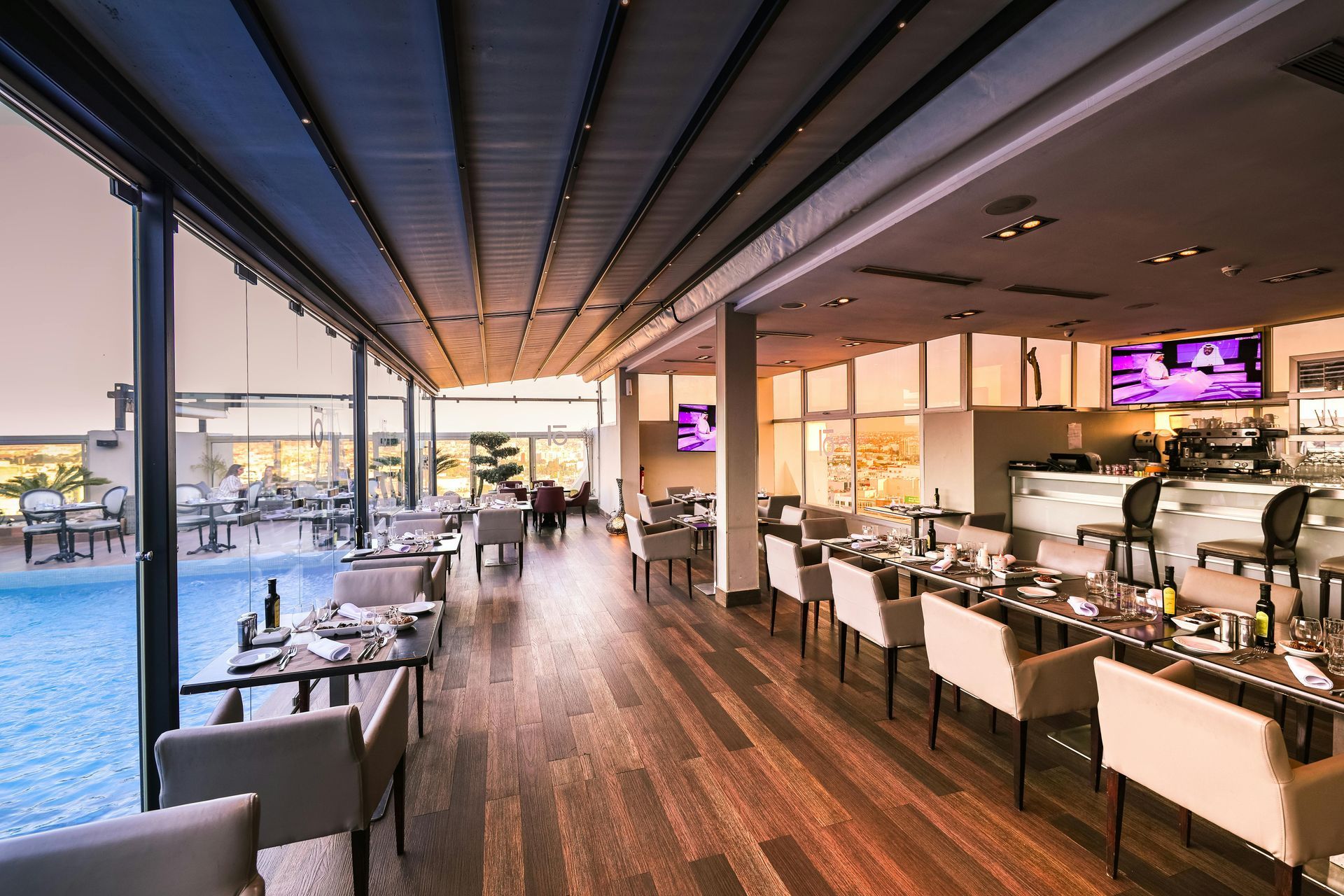 Restaurant interior with tables set, overlooking a pool. Features include wooden floors, retractable roof, and large windows.