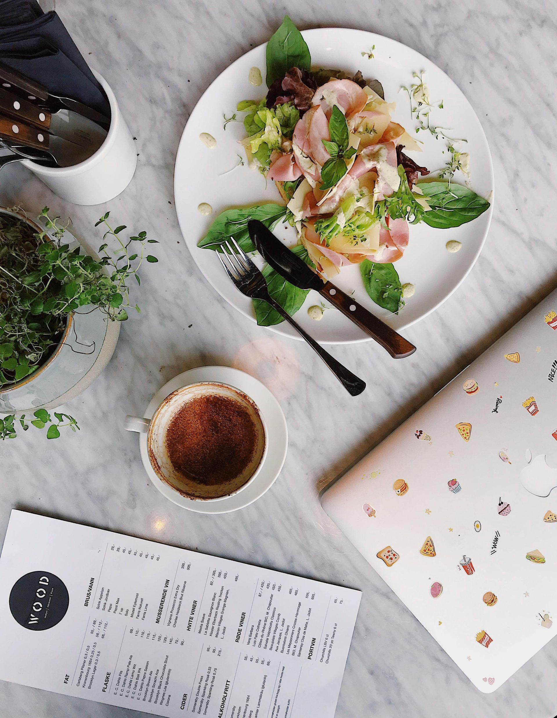 Brunch table with a plate of food, coffee, menu, laptop, and greenery.