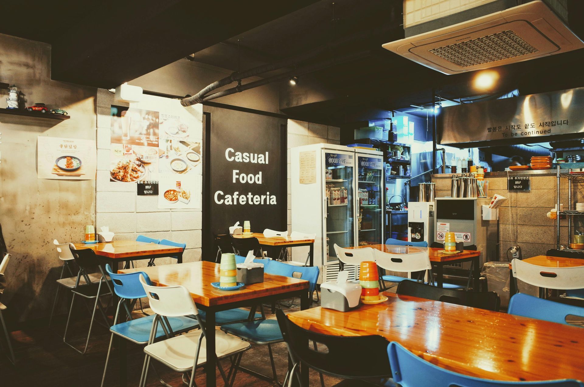 Interior of a casual food cafeteria with wooden tables, blue and white chairs, and a black chalkboard.