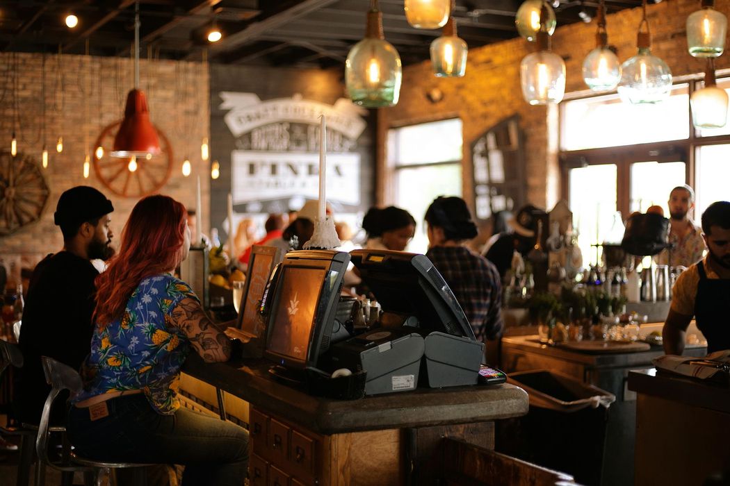 People at a bar, a woman with red hair sits at the counter, brick and metal accents.