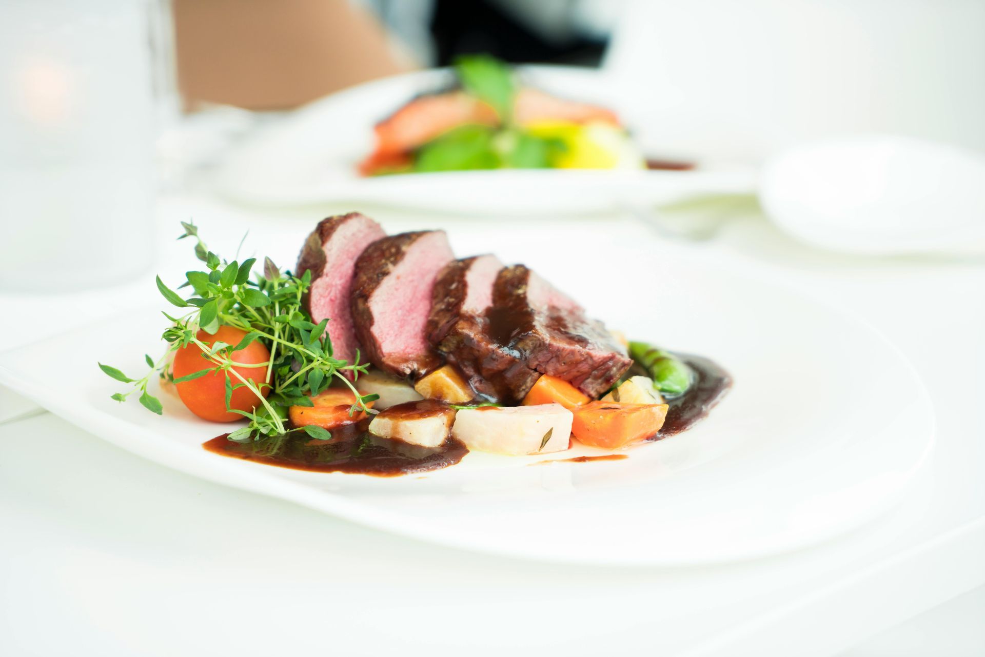 Sliced steak with sauce and vegetables on a white plate, fine dining setting.