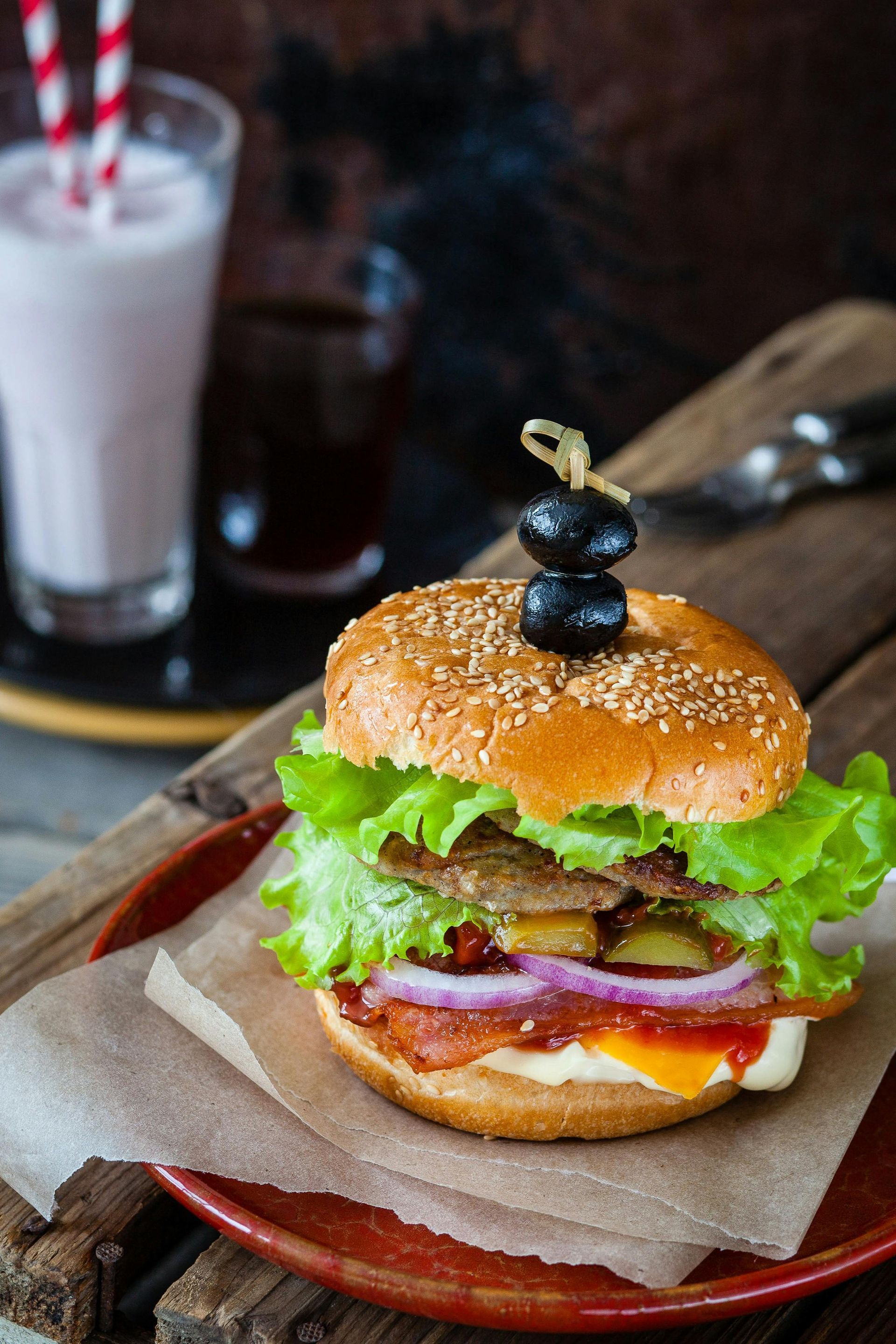 Burger with lettuce, tomato, and bacon on a plate with a shake and drink in the background.