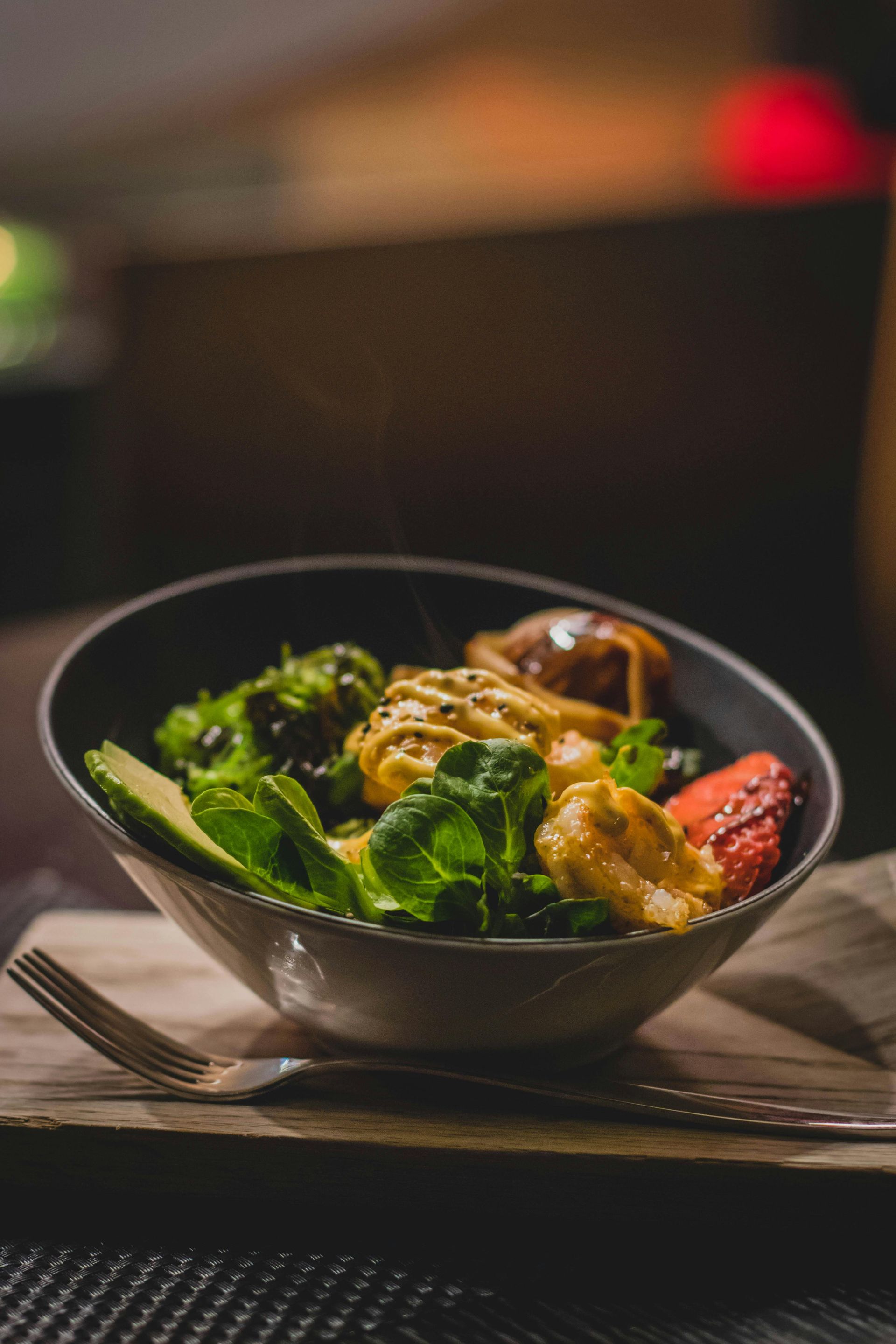 Salad bowl with avocado, greens, orange sauce, and a fork on a wooden board.