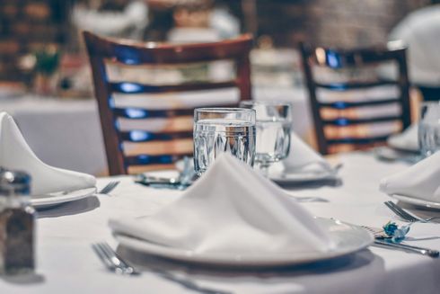 Table set for a formal meal with white tablecloth, napkins, glasses, silverware, and wooden chairs.