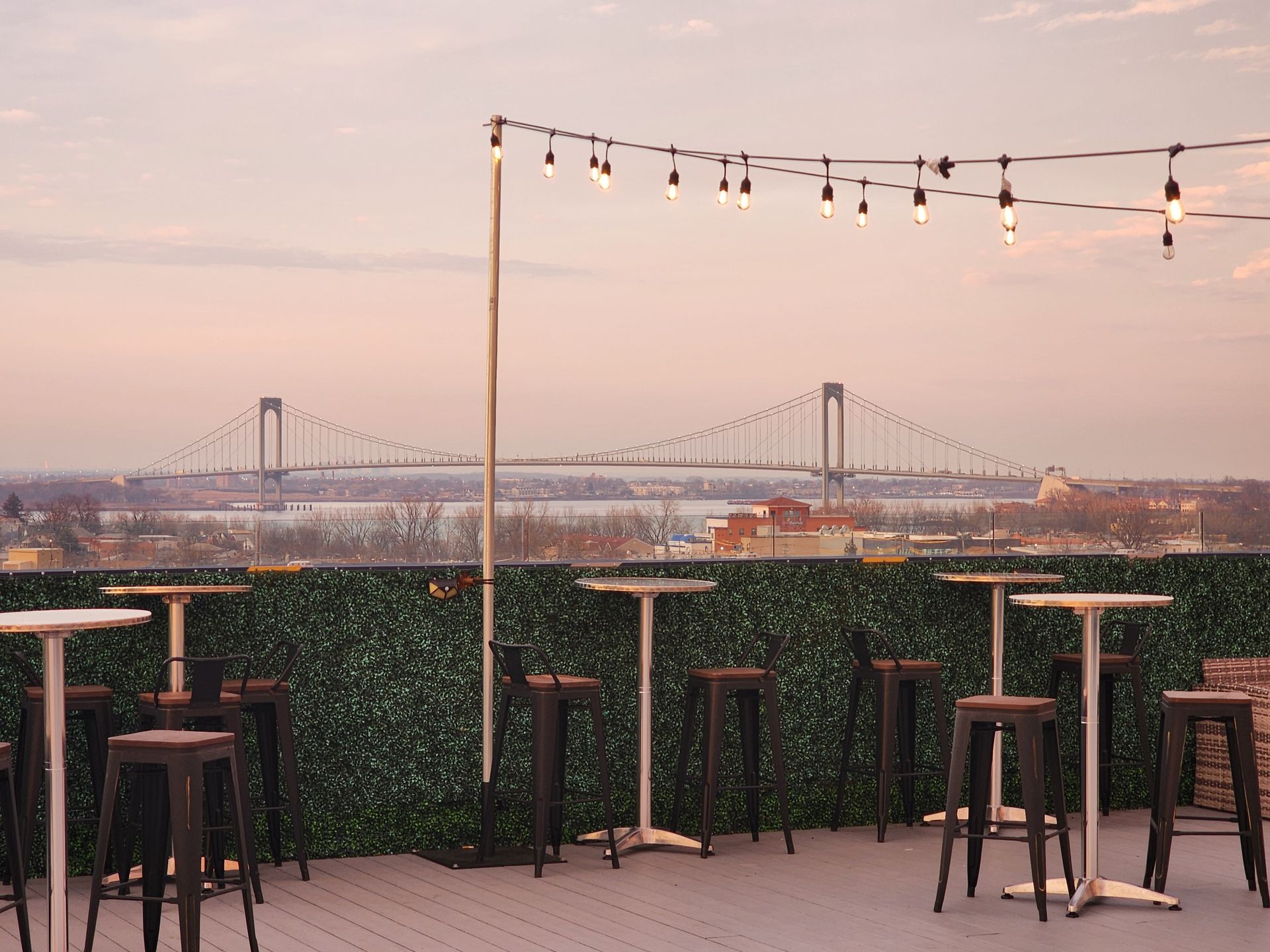 A row of tables and stools on a rooftop with a bridge in the background.