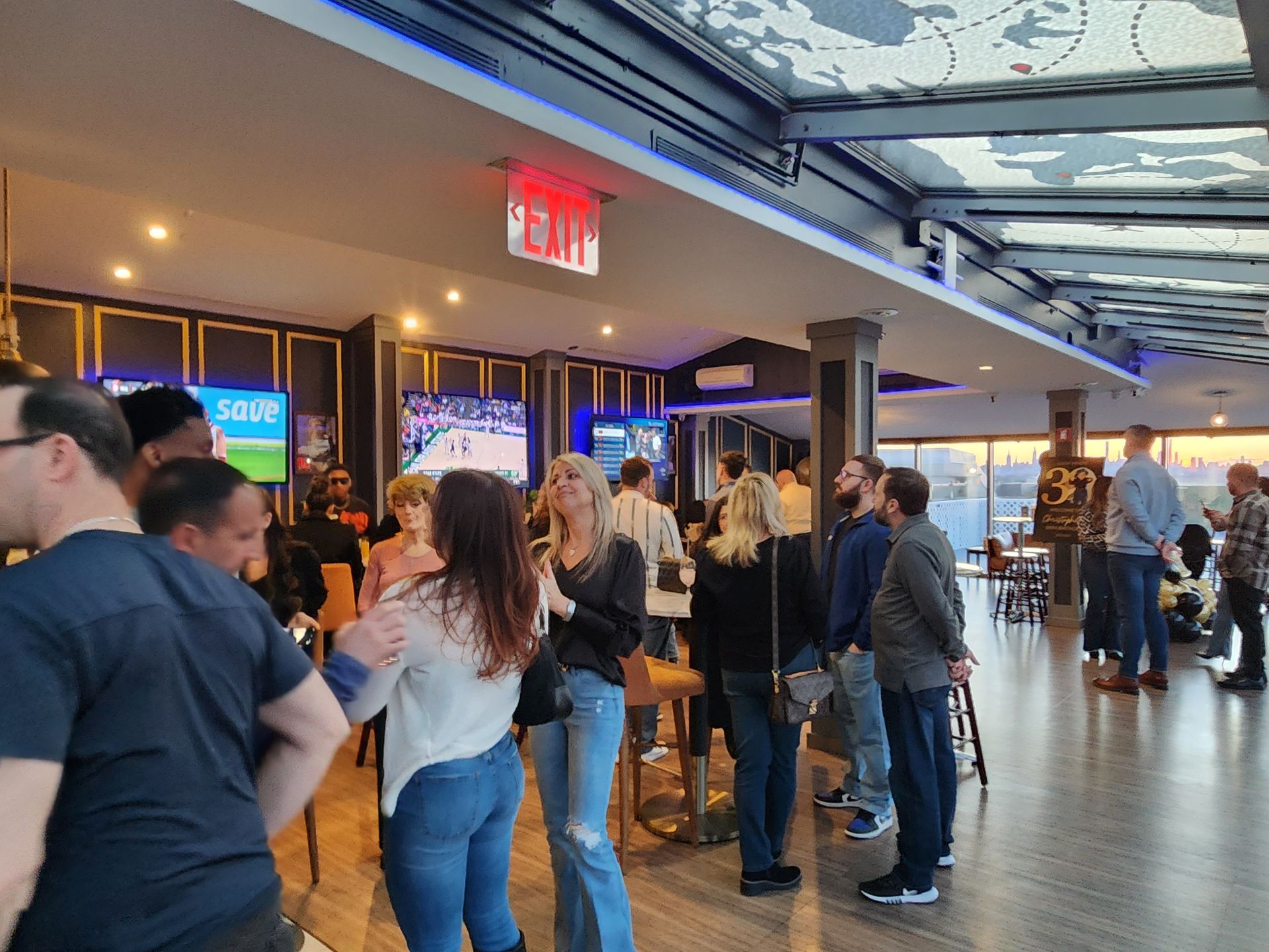 A group of people are standing in a room with a red exit sign on the ceiling.