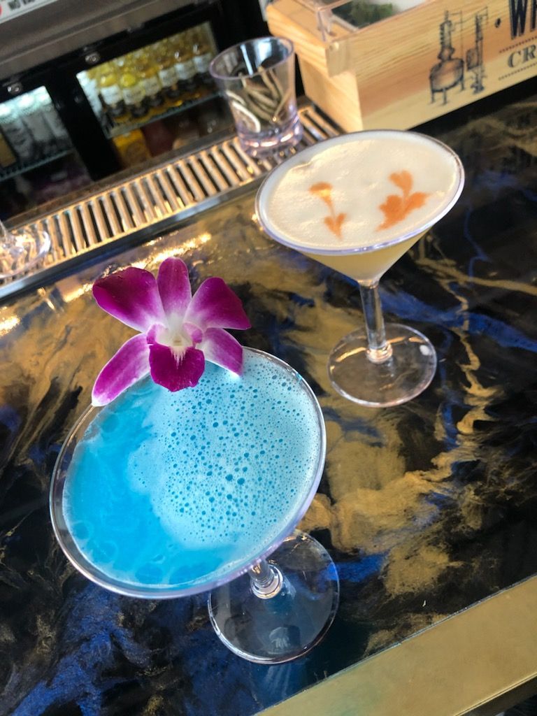 Two cocktails are sitting on a bar with a purple flower on top.