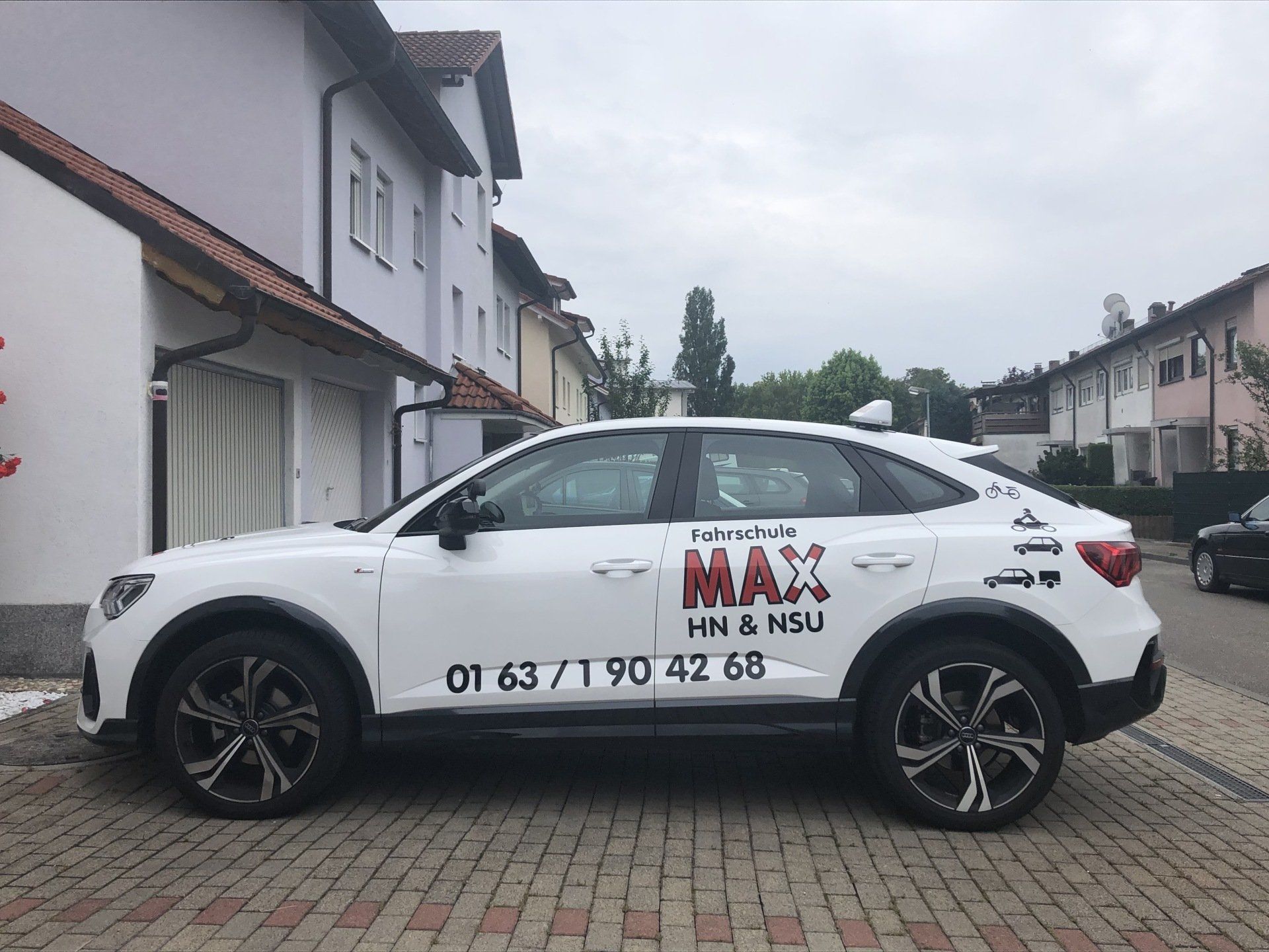 Ein weißes Auto mit dem Wort „Max“ auf der Seite steht vor einem Gebäude.