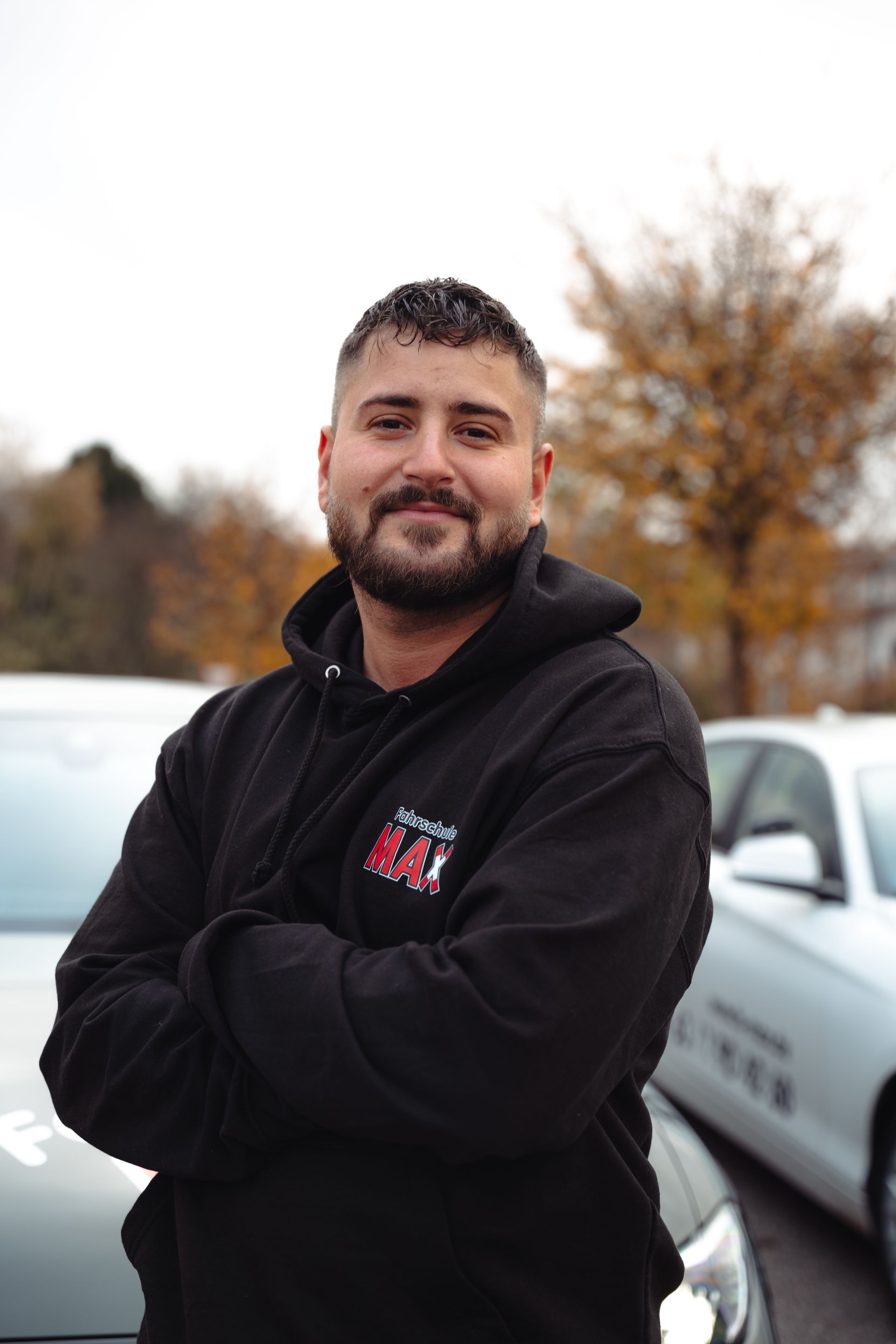 Ein Mann mit Bart steht mit verschränkten Armen vor einem Auto.