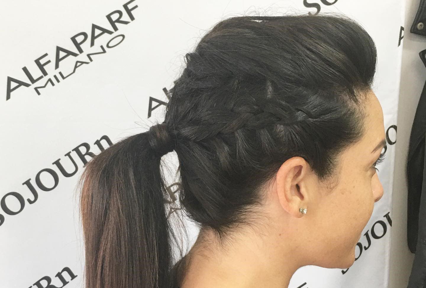 A woman wearing a braided ponytail is standing in front of a wall that says alfaparf milano