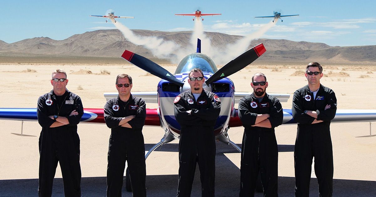 Five pilots standing in front of a plane from Sky Combat Ace. Three Sky Combat Ace planes are flying in the background.