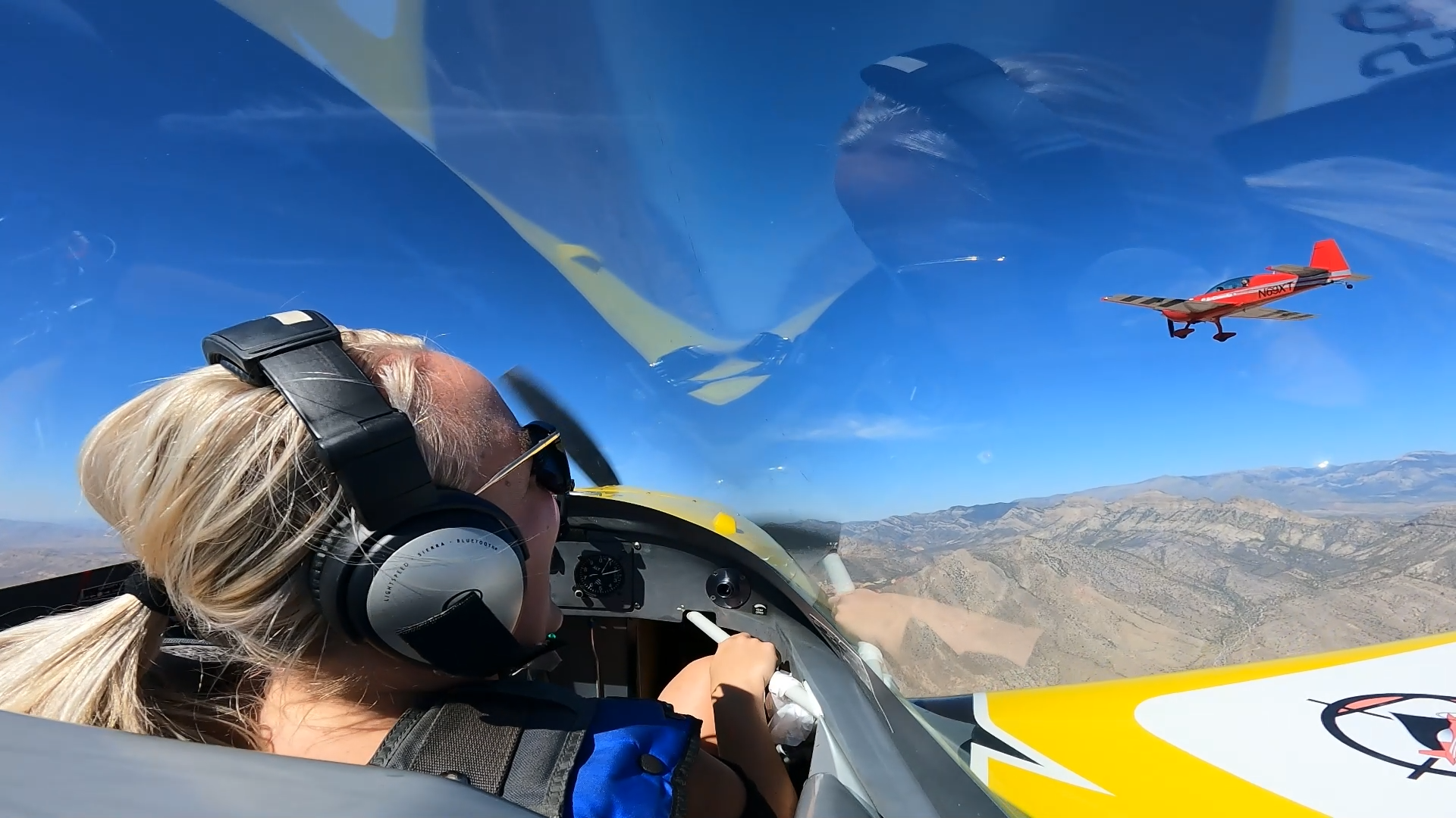 A pilot in a cockpit looks toward another small red airplane flying nearby over a mountainous landscape.