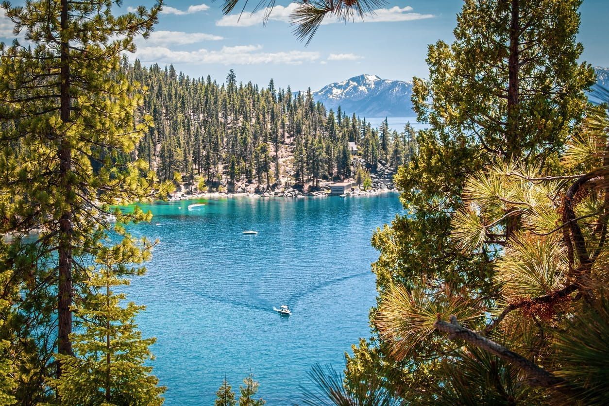 An aerial view of Lake Tahoe with a boat cruising through the lake near the forest. Mountains appear in the background.