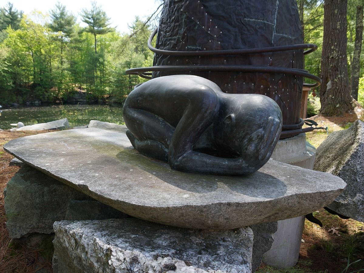 Bronze sculpture of a figure curled in a fetal position on a stone slab, set outdoors with a water feature behind.