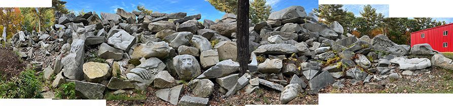 A long pile of gray stones in front of some trees and a red building under a blue sky.