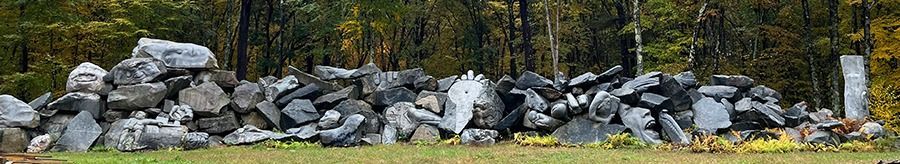 A long, low stone wall stretches across a grassy area in front of a treeline. The wall is made of various sized rocks and boulders.