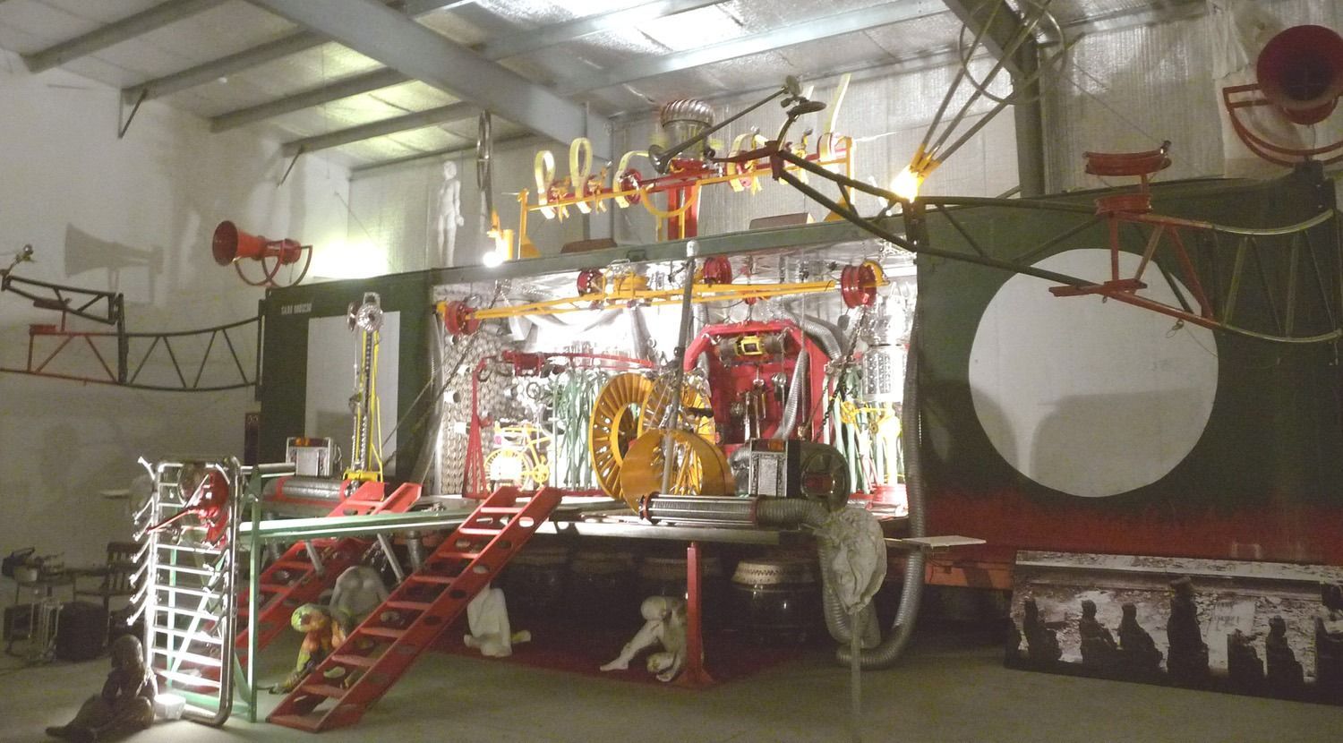 Installation art piece inside a white-walled space. The artwork incorporates red, gold, and silver mechanical-looking components, against a backdrop with a green circle and red border.