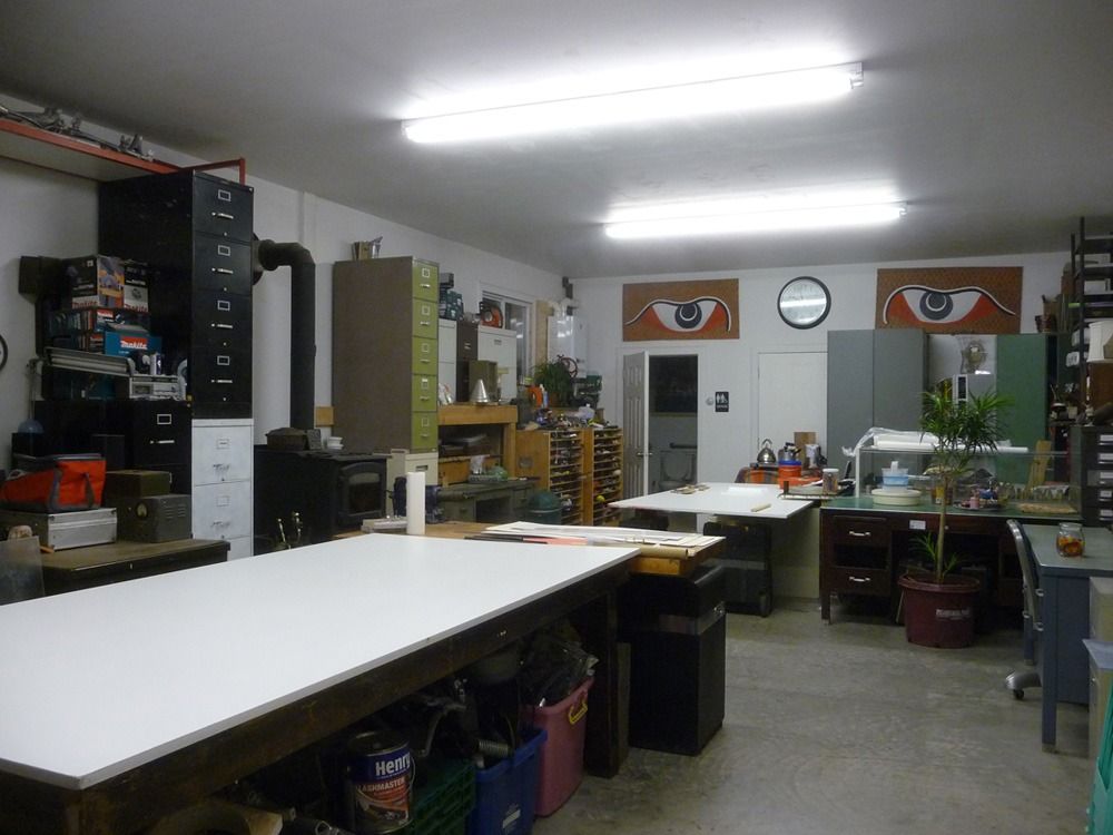 A cluttered workshop with large worktables, filing cabinets, and artwork on the walls. Fluorescent lights illuminate the space.
