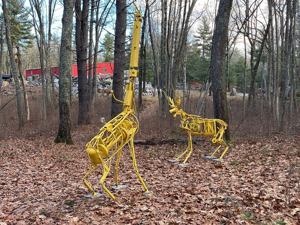 Two yellow metal sculptures of a giraffe and deer in a wooded area with fallen leaves.