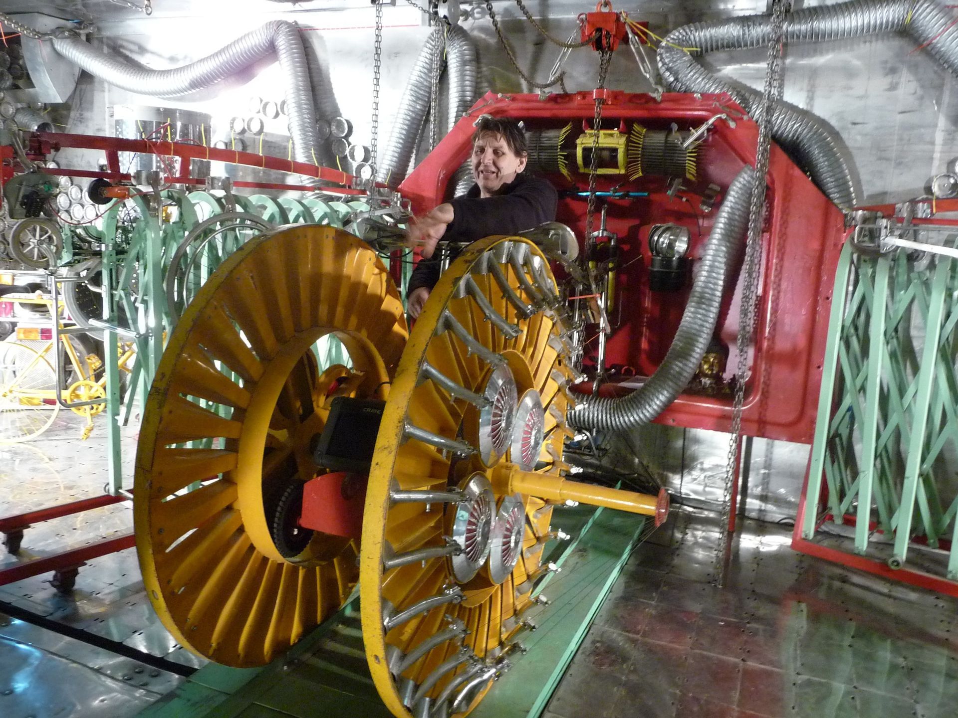 A person in a complex machine with large, yellow spiral wheels. Red machinery and metal pipes fill the background.