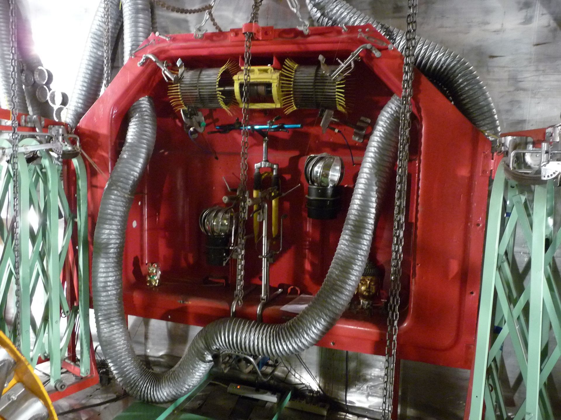 Red, boxy machinery with gray corrugated hoses, suspended inside a metal frame, likely industrial equipment.