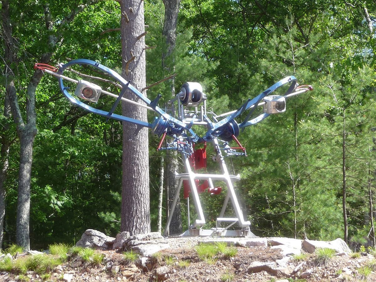 A metal sculpture resembling a bird with outstretched wings, placed in a wooded area.