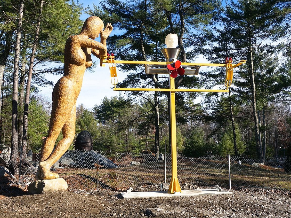 Golden statue of a woman looking toward a yellow metal contraption in a wooded area.