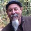 Mature man with a white goatee, wearing a hat and coat, smiles outdoors in a forest setting.