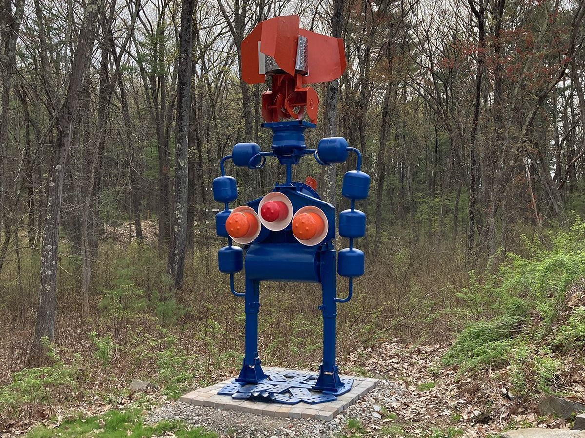 Blue metal robot sculpture with orange and red accents, standing on a concrete base in a wooded area.