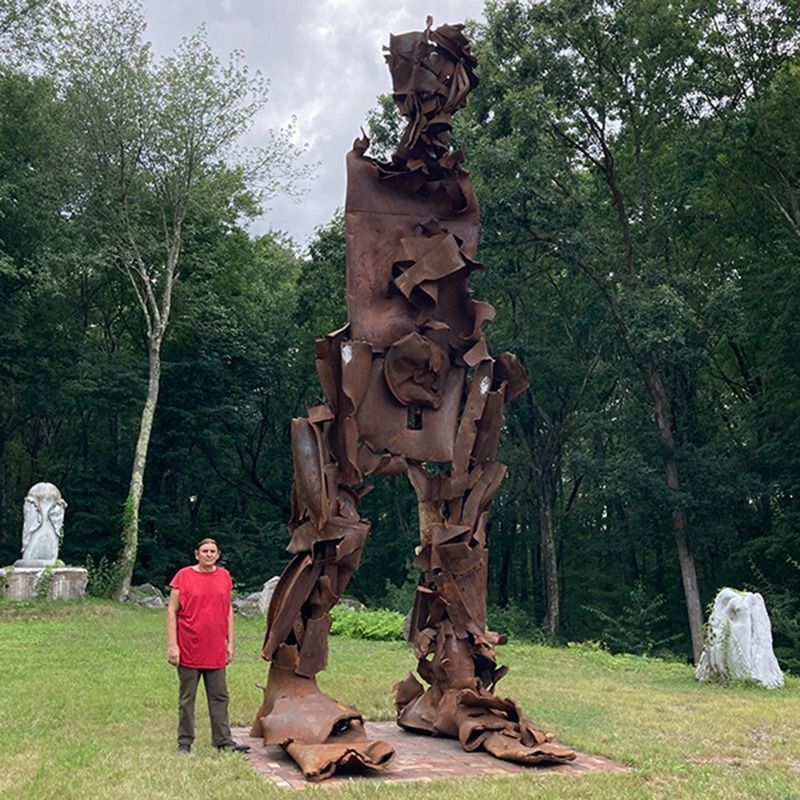 Two abstract, light-colored sculptures sit on wooden stumps in a sunlit forest clearing. Green foliage surrounds them.