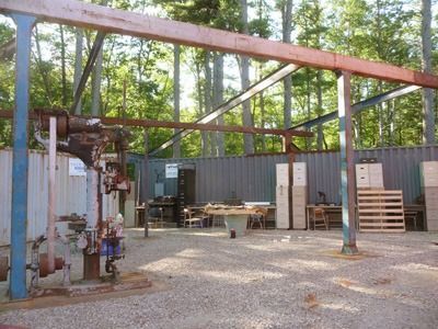 An outdoor workshop with a metal frame, a shipping container, and various tools and materials. Trees form the background.