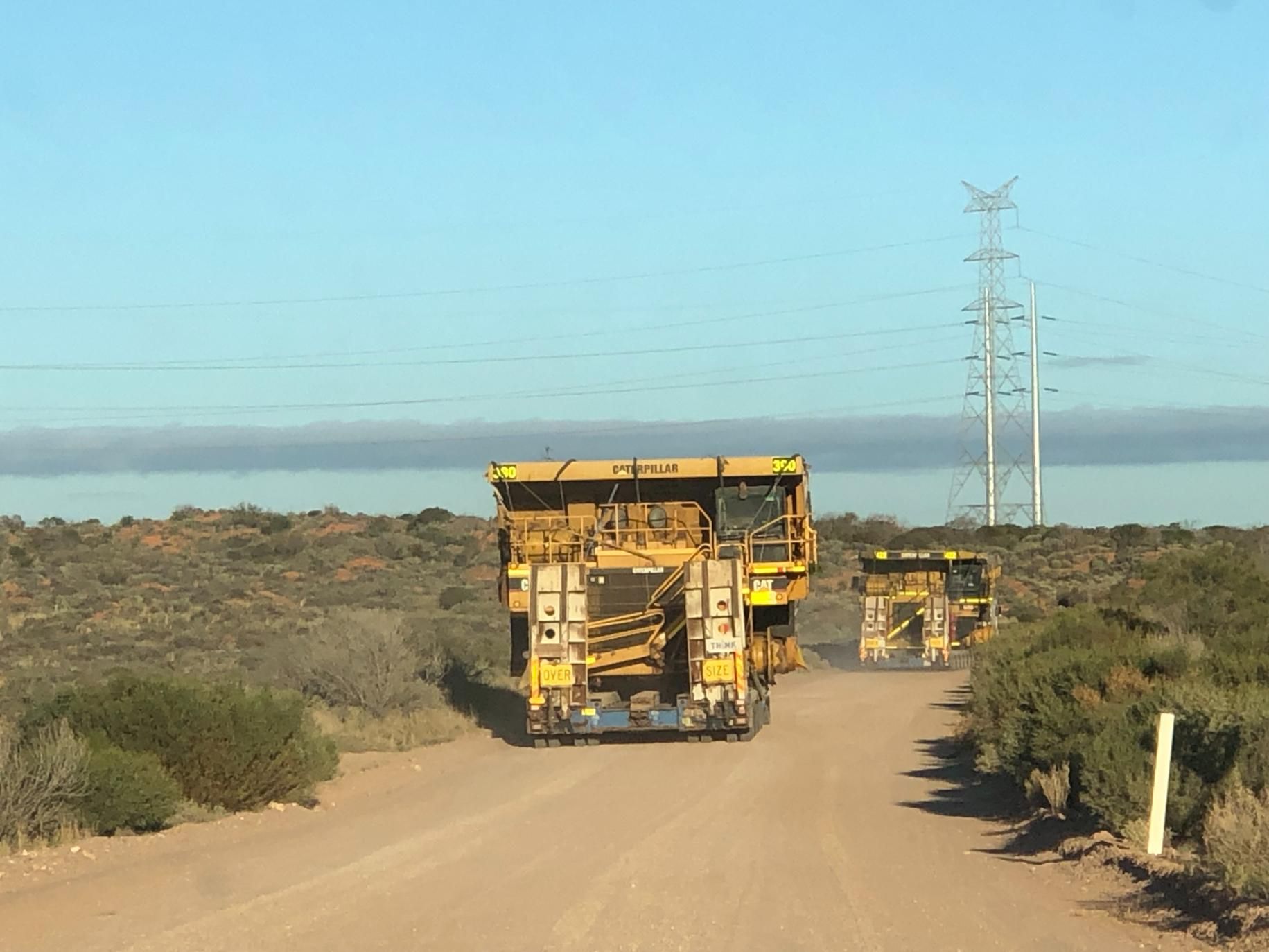 A Row of Yellow Trucks Are Driving Down a Dirt Road — Col Adamson Transport Pilot Services In Farleigh, QLD