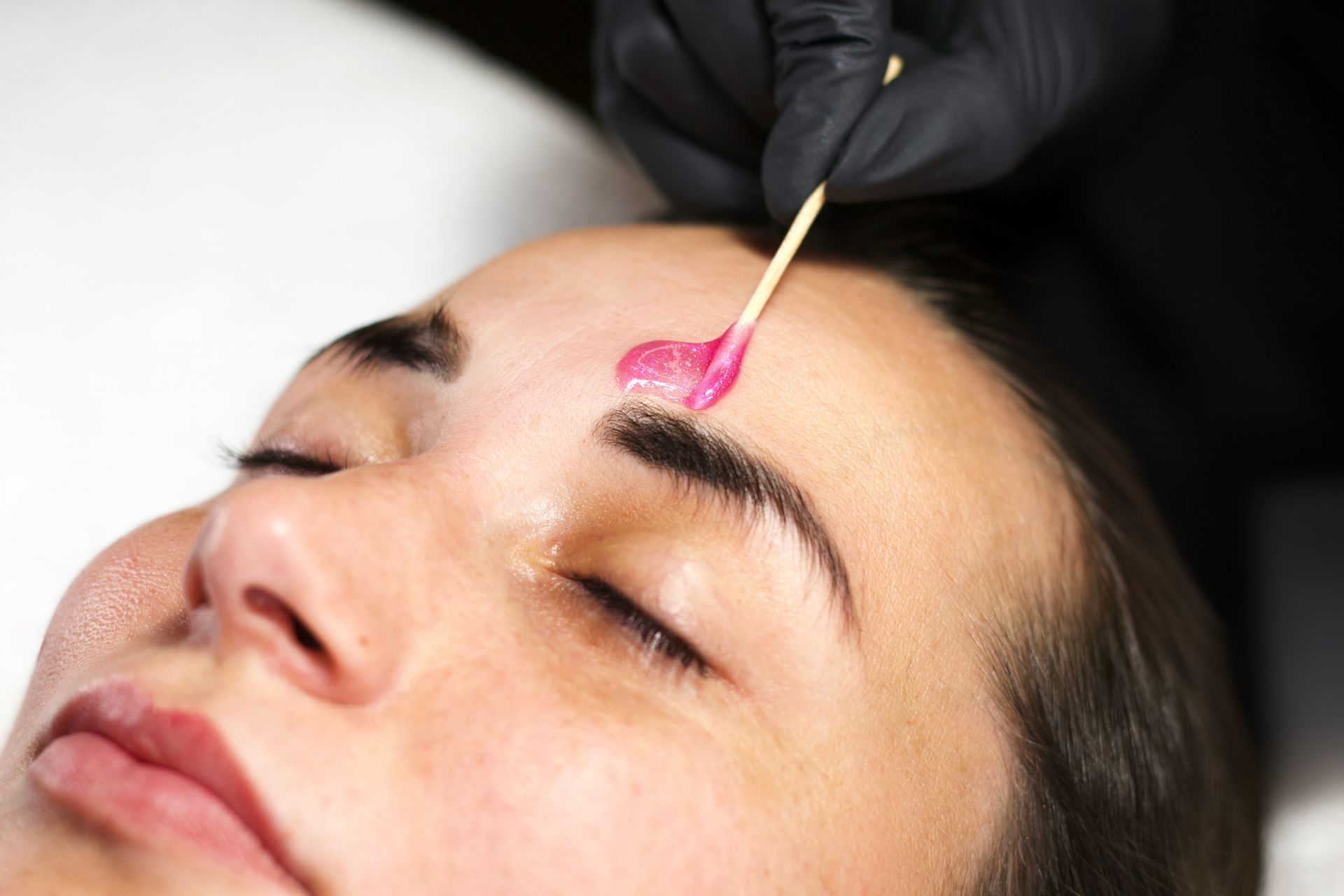 Waxing eyebrows. Person's face, eyes closed. Gloved hand applies pink wax to brow with a stick.