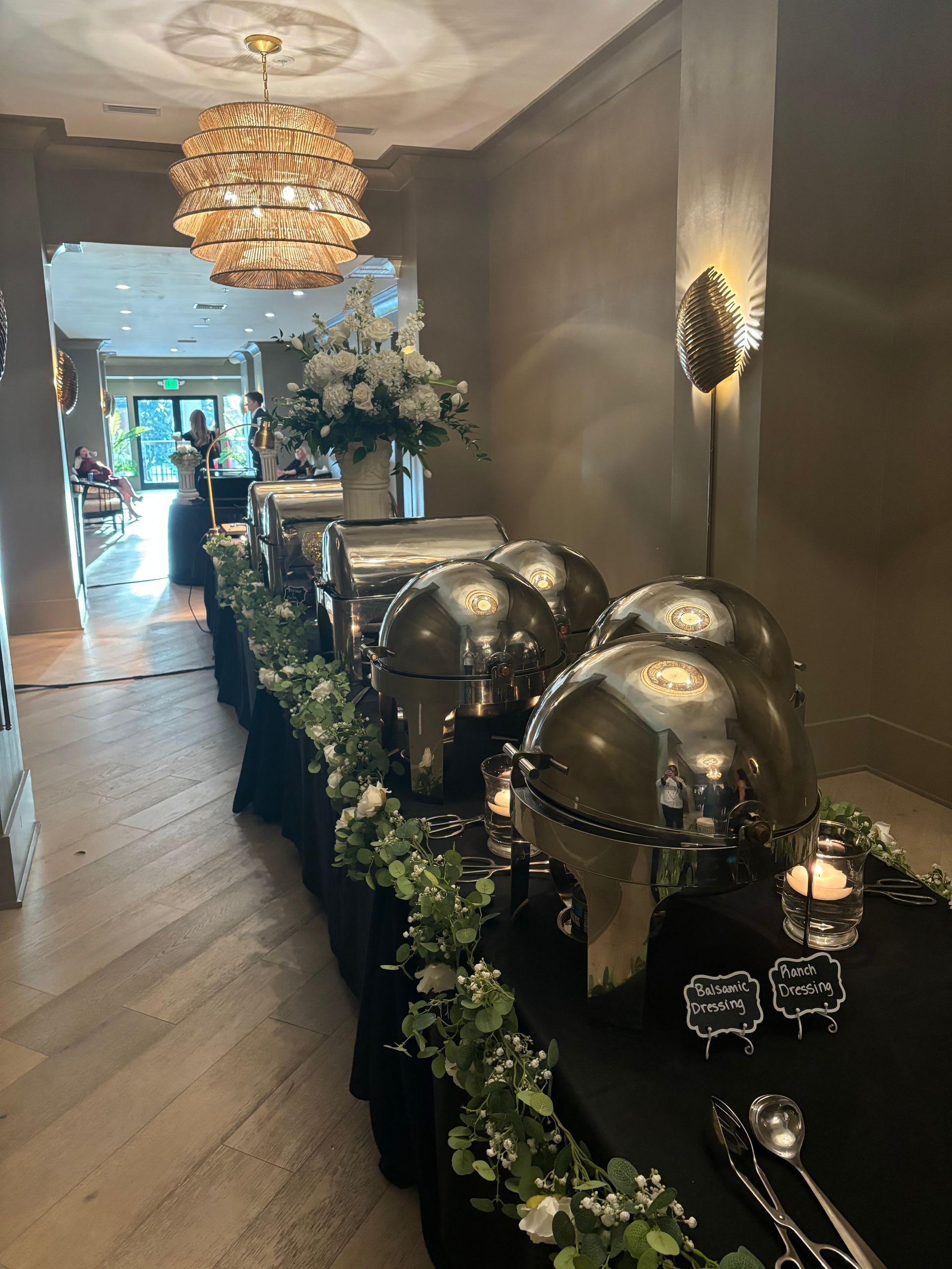 Buffet table with covered silver serving dishes, adorned with greenery and white floral arrangements, in a long hallway with a chandelier.
