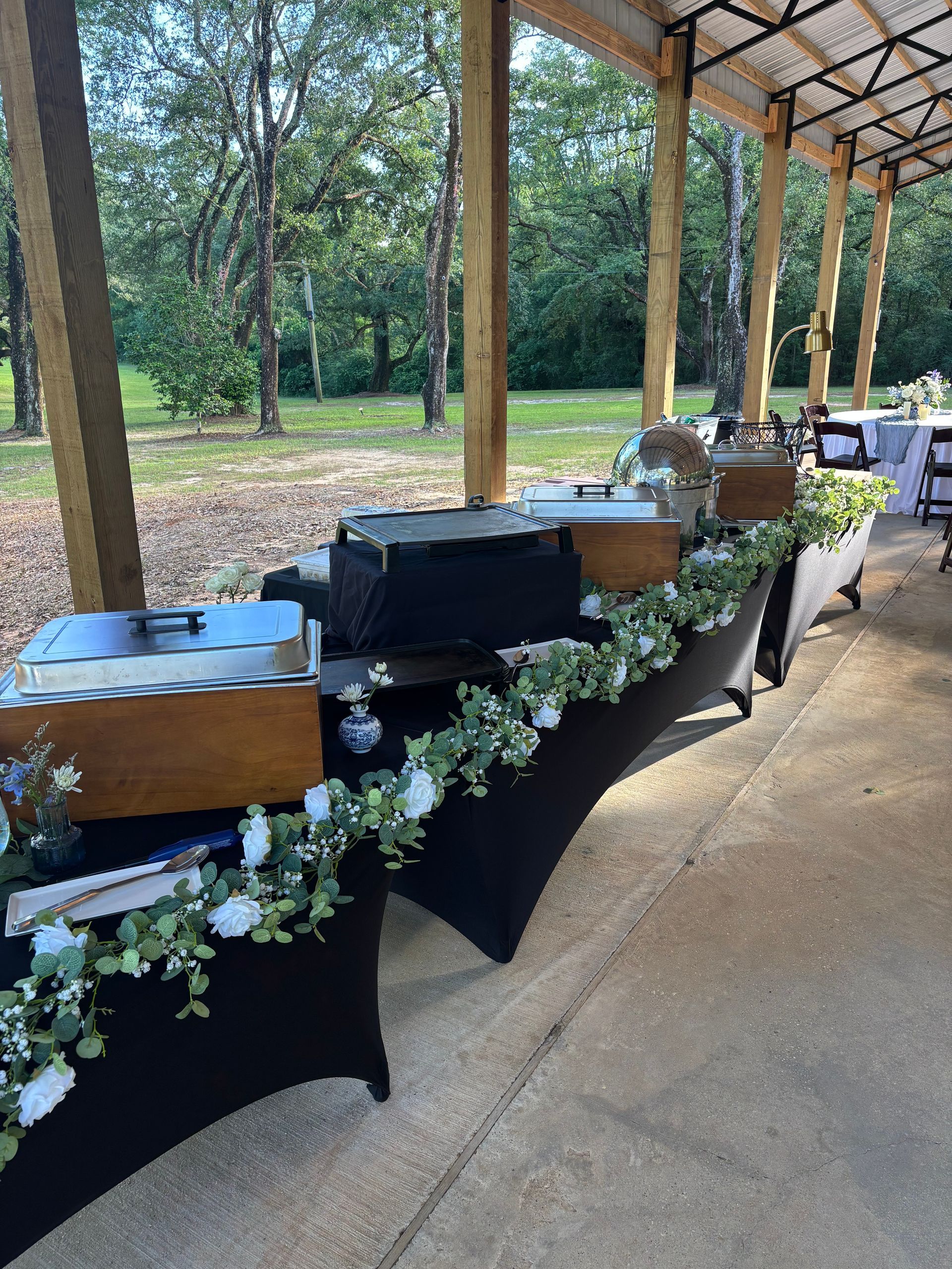 Buffet table at an outdoor event, draped in black with food warmers. A garland of white flowers and greenery decorates the front.