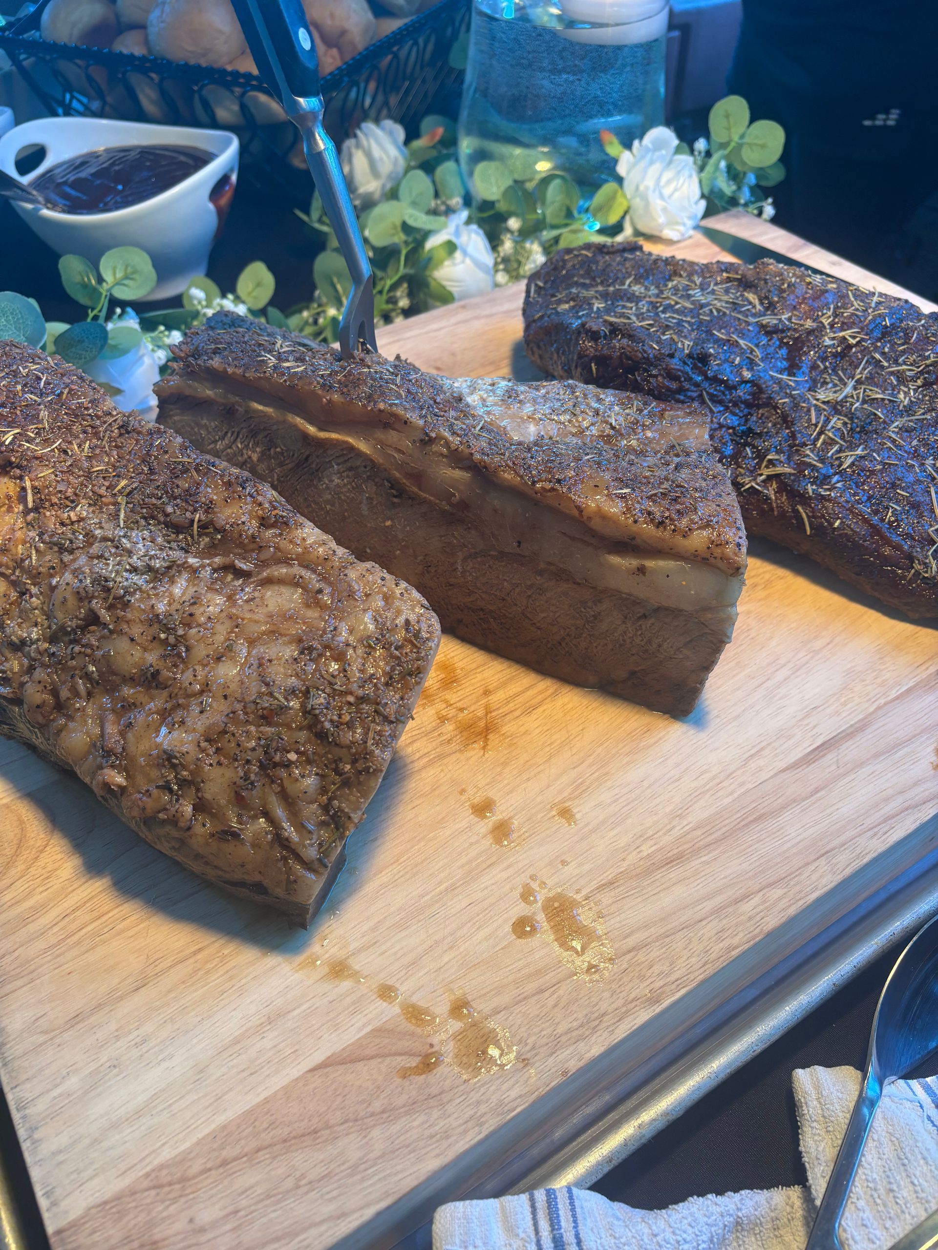 Four seasoned, cooked meat cuts on a wooden board, with a dipping sauce and flowers in the background.