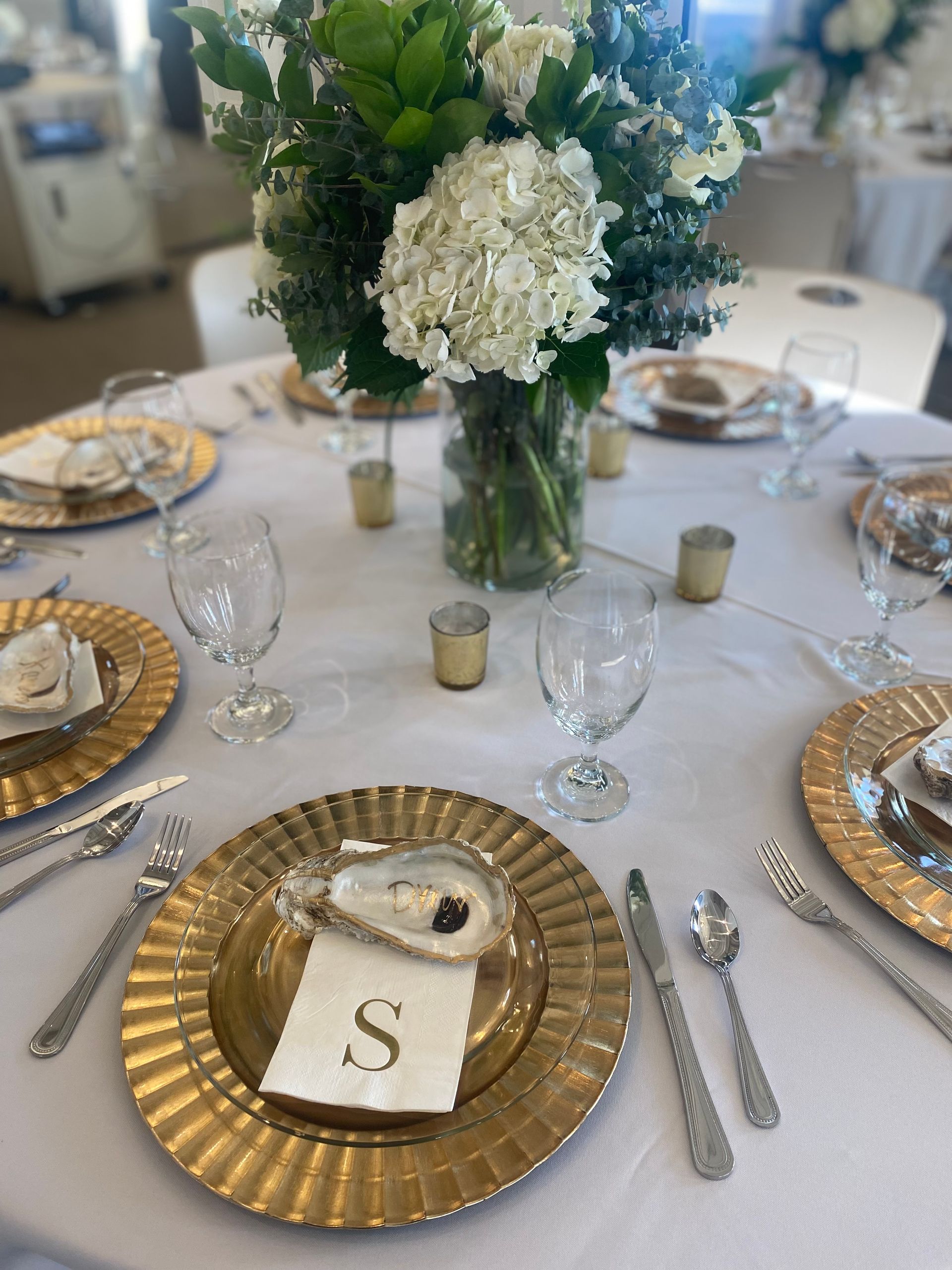 Wedding table setting with gold chargers, oyster shells, and a floral centerpiece. White tablecloth, clear glassware, and personalized place cards.