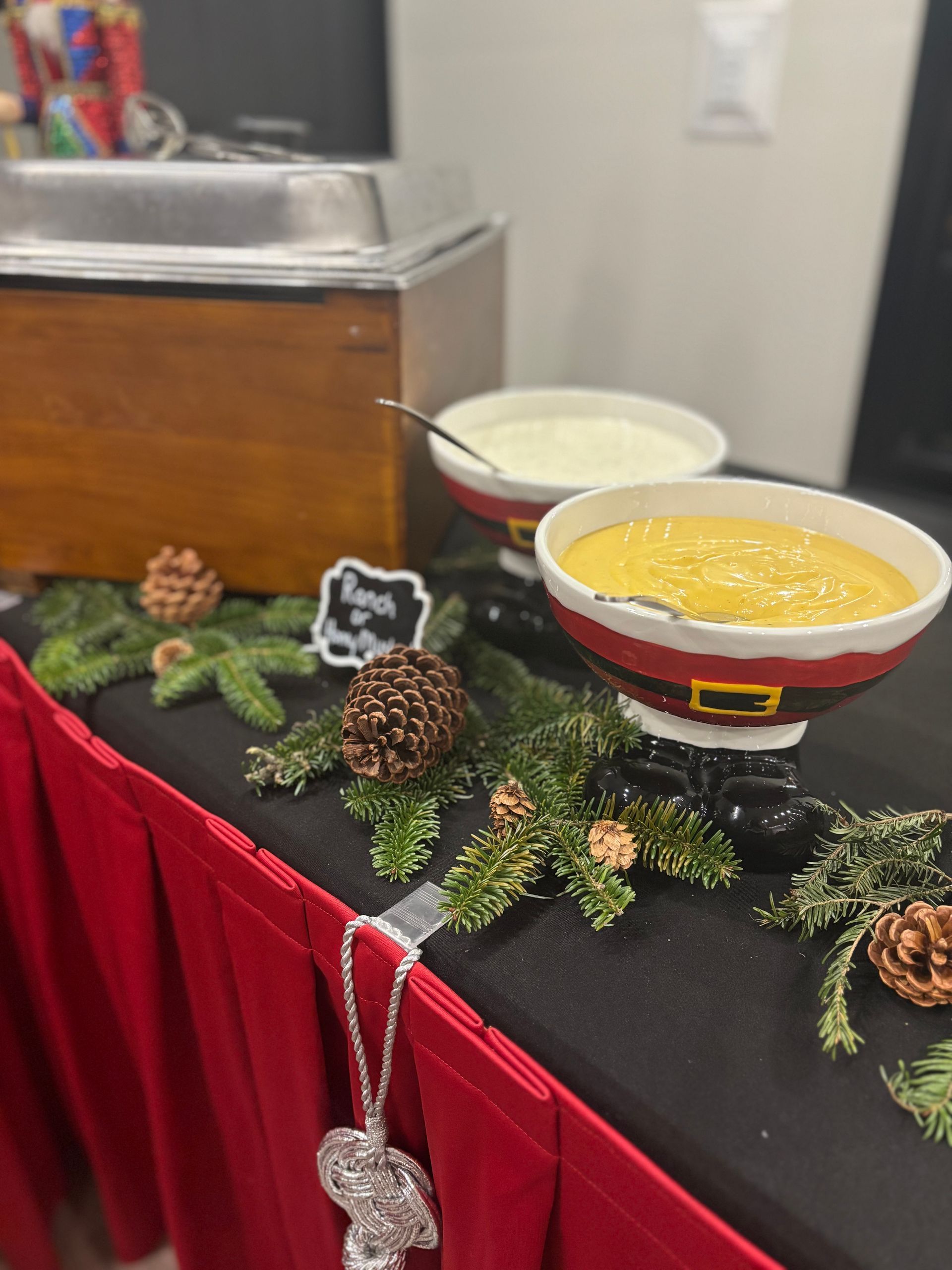 A festive food display: two bowls of dip in Santa-themed containers, surrounded by greenery and pinecones, on a black table with a red skirt.