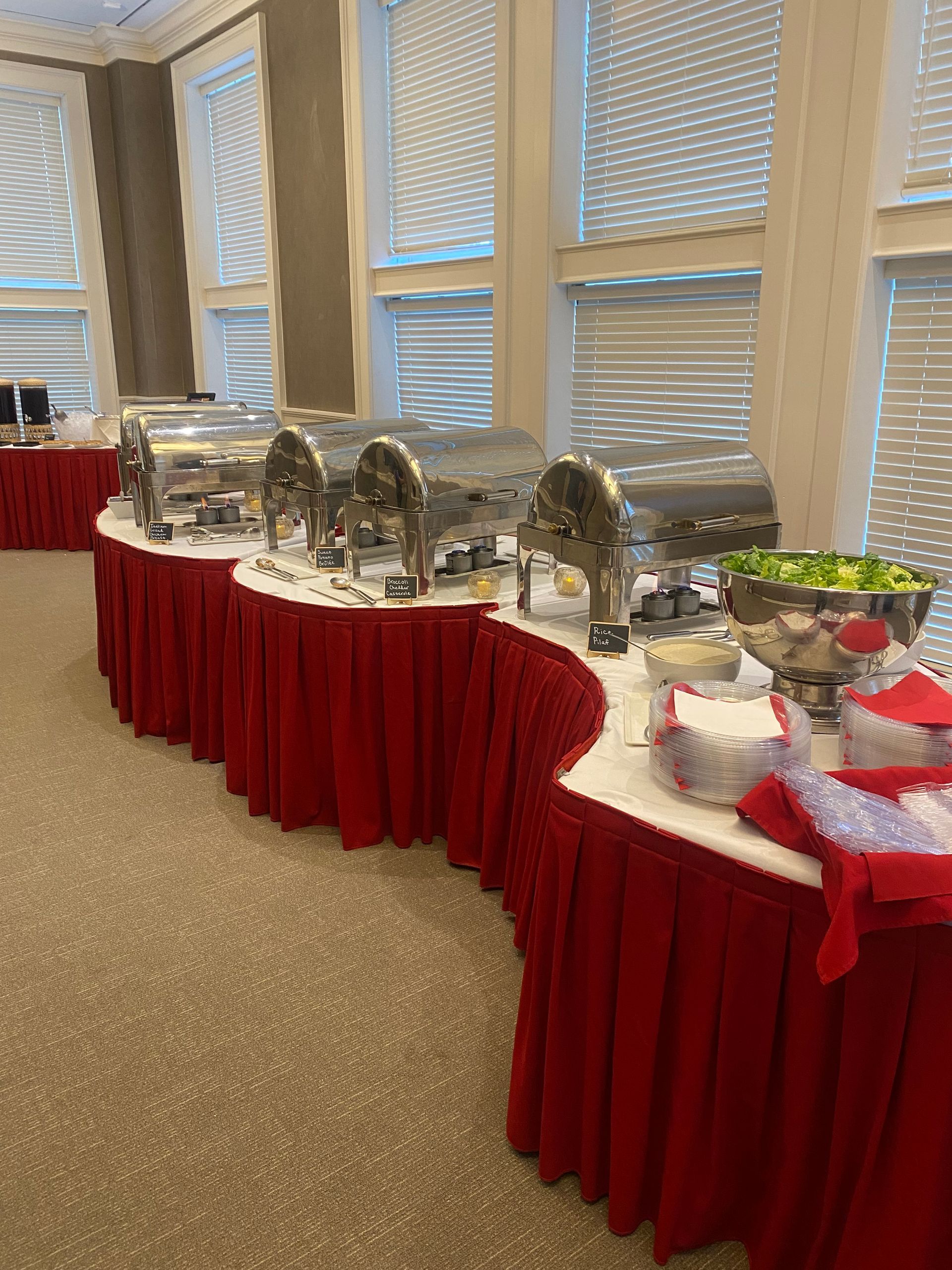A buffet table draped in red cloth holds metal food warmers. A room with large windows provides the background.