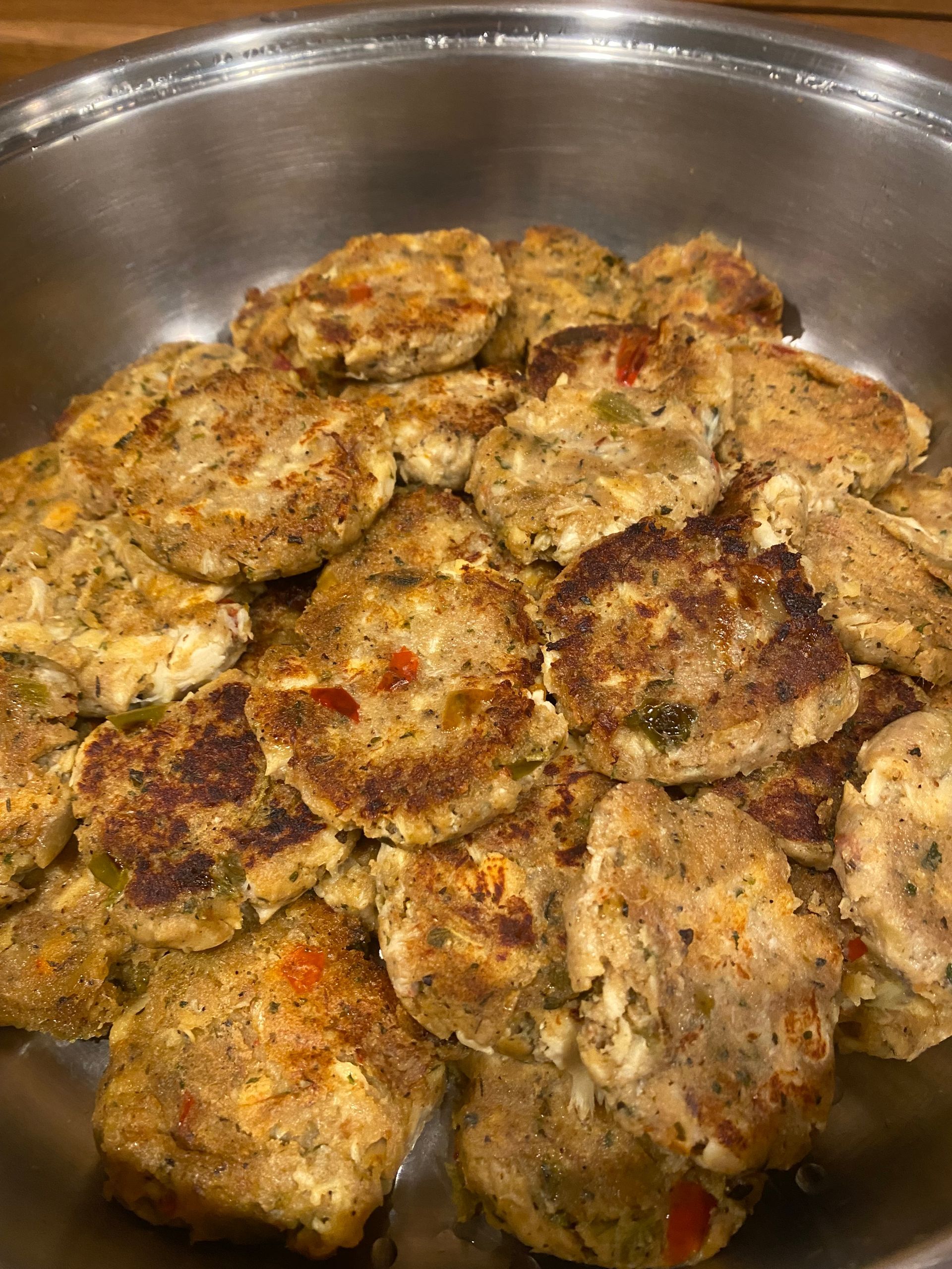 A metal bowl filled with many cooked crab cakes; golden brown with visible vegetables.