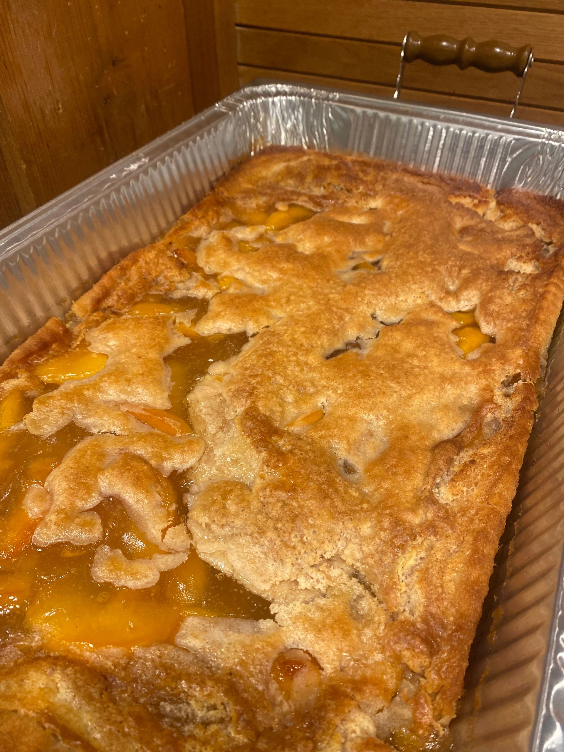 Peach cobbler in a foil pan; golden crust with visible peach slices and golden, syrupy juices.