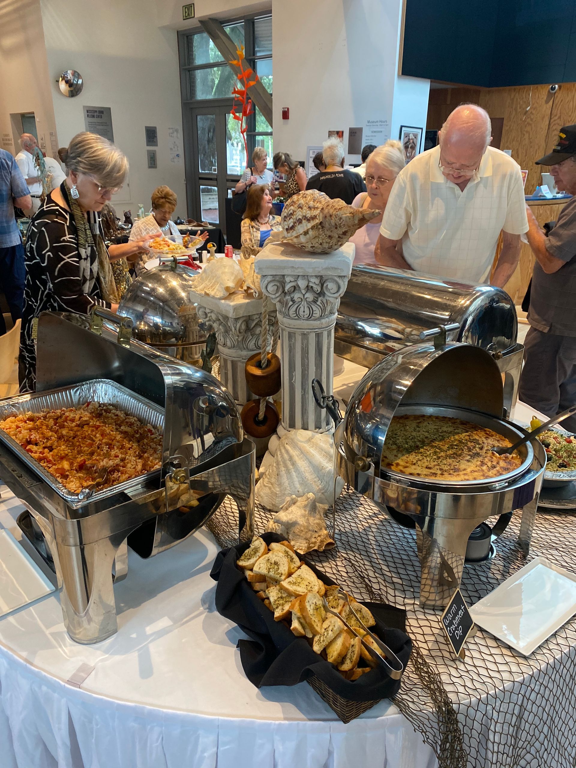 People at a buffet with various dishes in silver serving containers. A decorative pillar and shells are also displayed.
