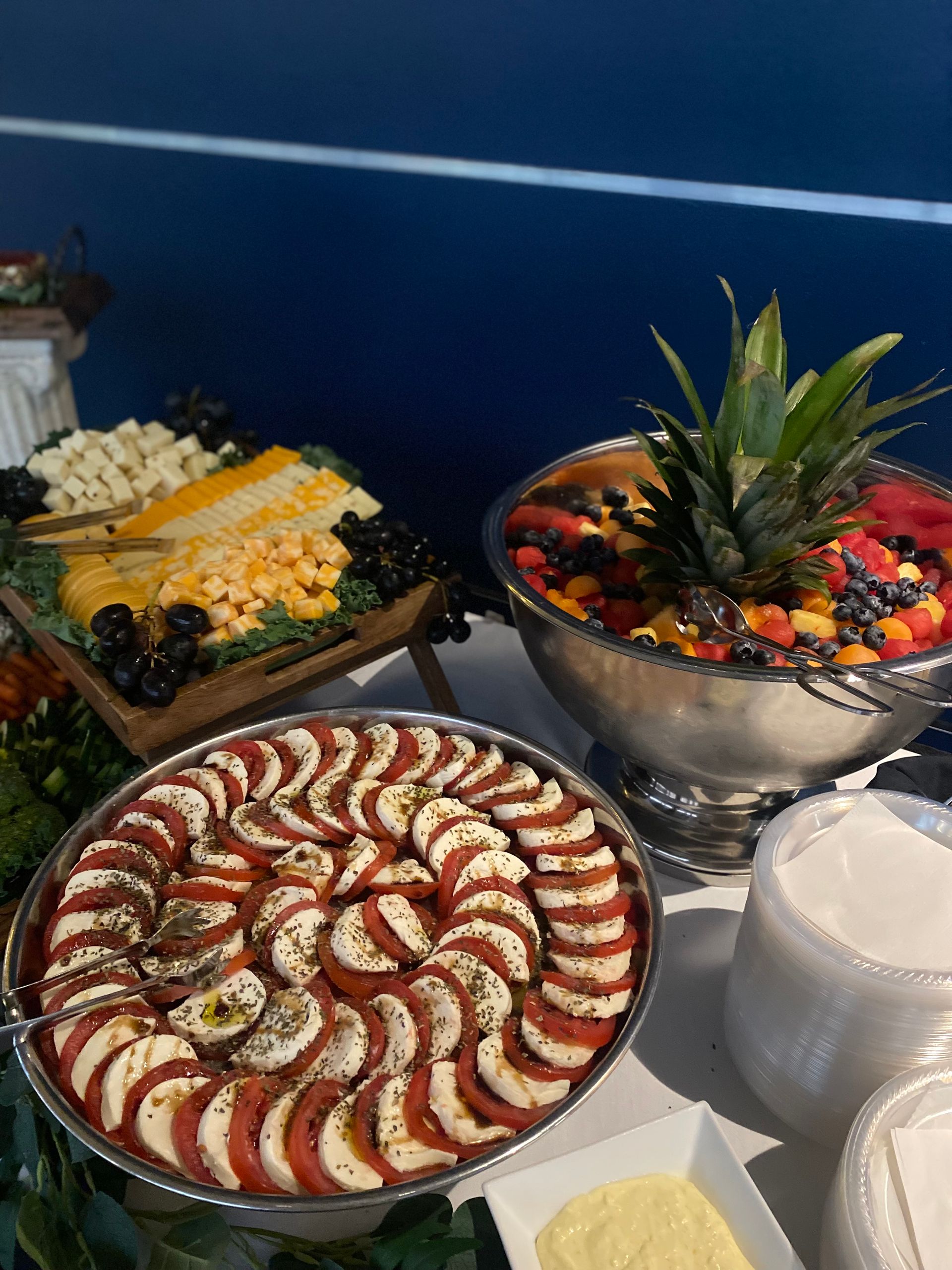 A buffet table with a caprese salad, cheese platter, and fruit arrangement. The fruit arrangement has a pineapple top.