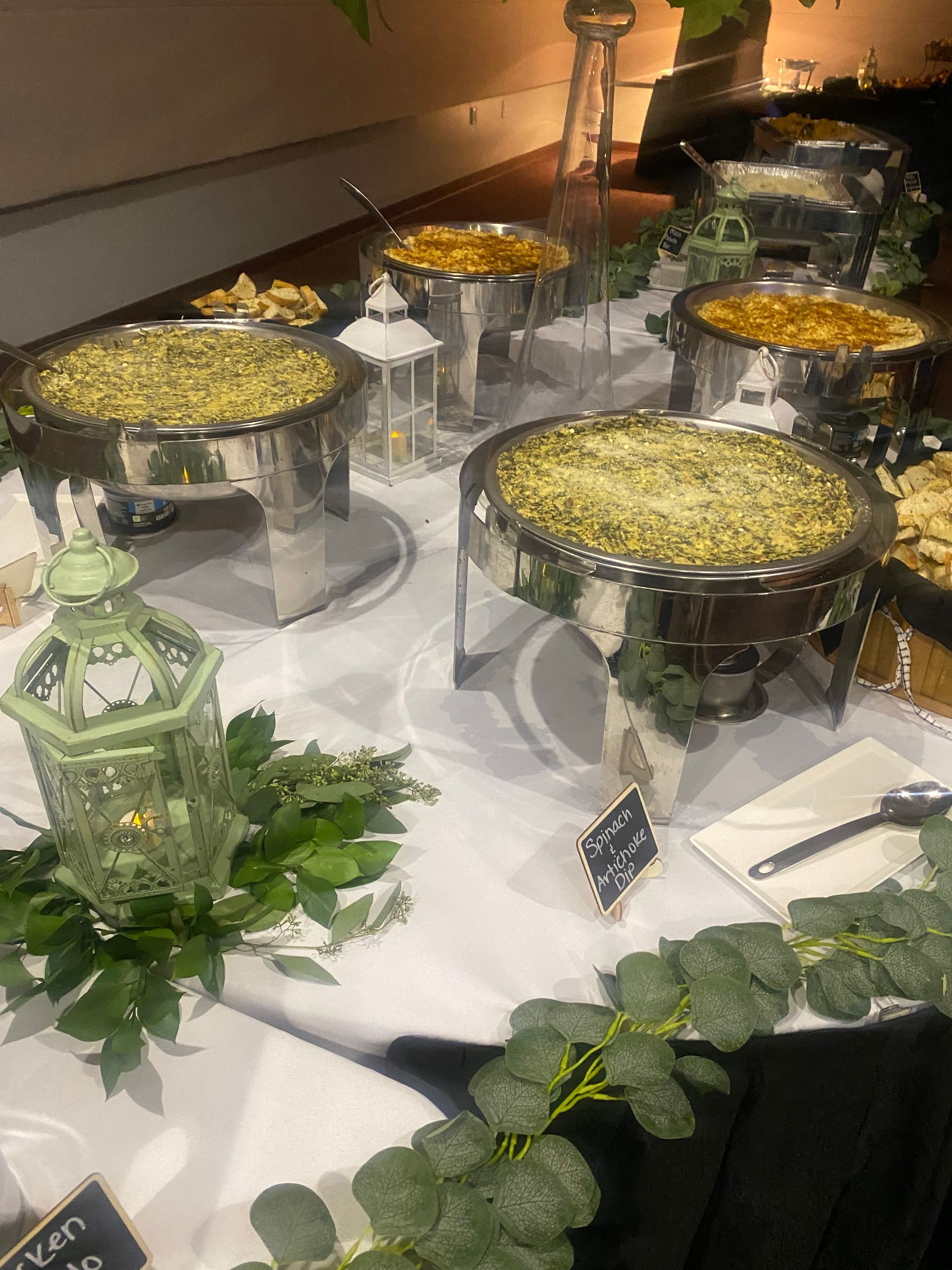 Buffet table with several chafing dishes containing food. Greenery and decorative lanterns surround the table.